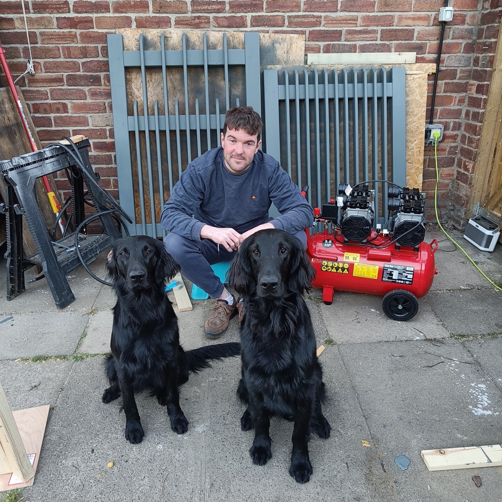 An image of the business owner sat in front of two gates that he has been spray painting. There are two black flatcoat retrievers sitting in front of the man looking at the camera