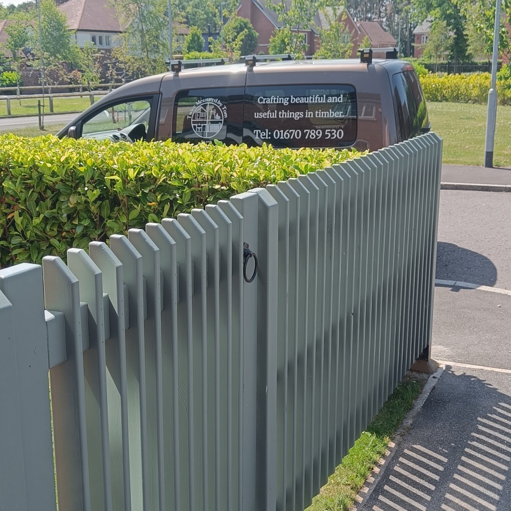 A picket fence and side gate for a front garden. Pickets are deep and rebated so that the fence rails sit within the depth of the pickets. The rest of the fence and gate are painted in Farrow & Ball Green Smoke. There is a hedge and branded van parked beyond the fence.