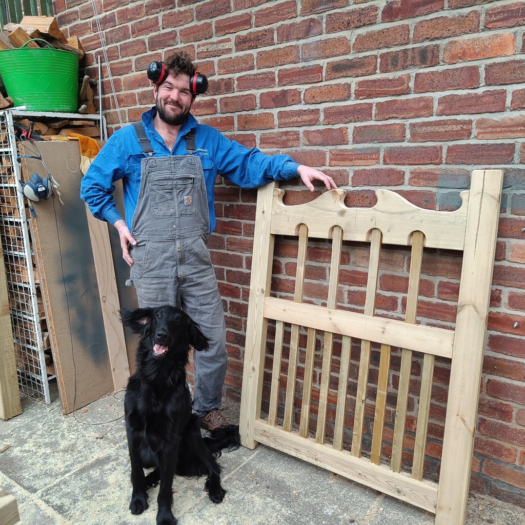 Man and black dog standing next to hand made wooden gate with a distinctive wave pattern on the top bar and picket infills.